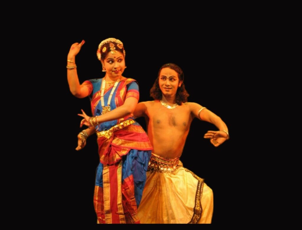 A male and female dancer performing a duet in traditional Indian classical dance attire, showcasing graceful expressions and hand gestures against a black background.